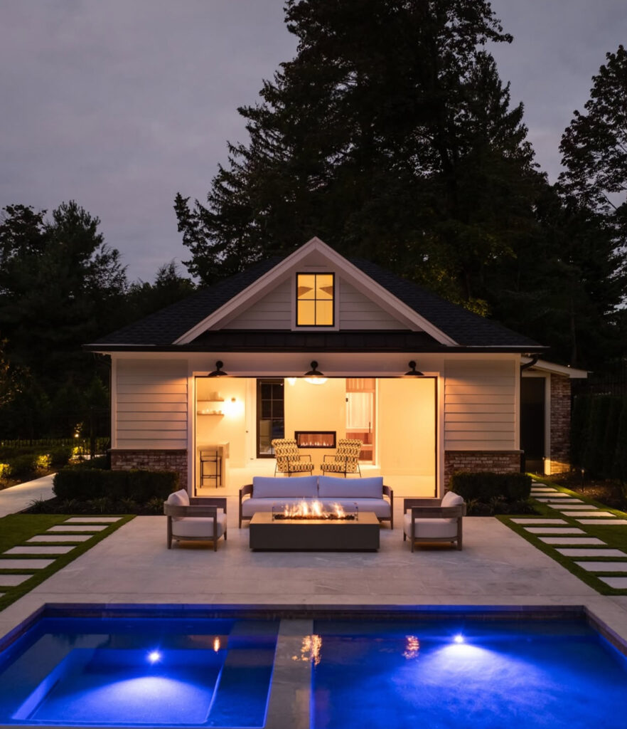 Illuminated pool and fire table facing the pool house at dusk