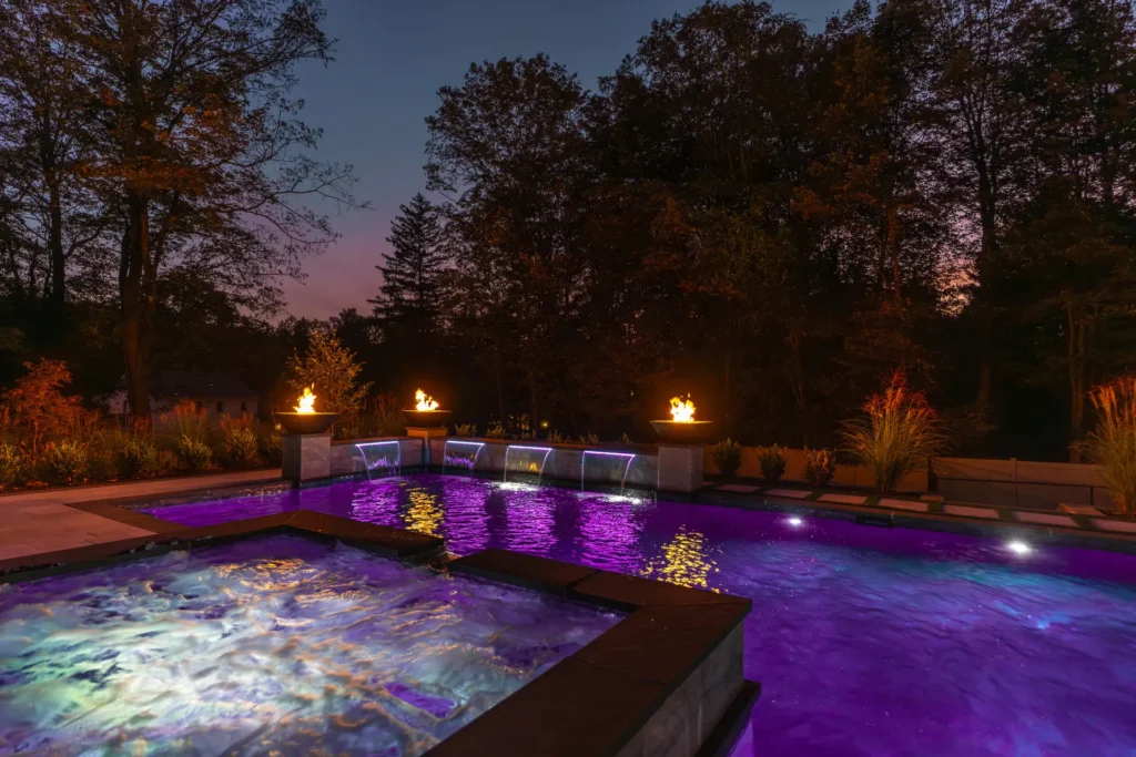 Pool and raised spa illuminated purple beneath fire bowls and waterfalls at dusk
