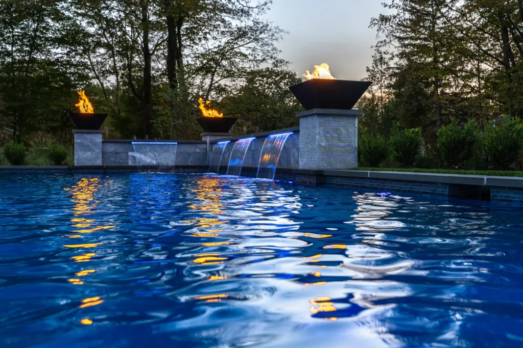 Close view across a blue-lit pool toward waterfalls and three fire bowls