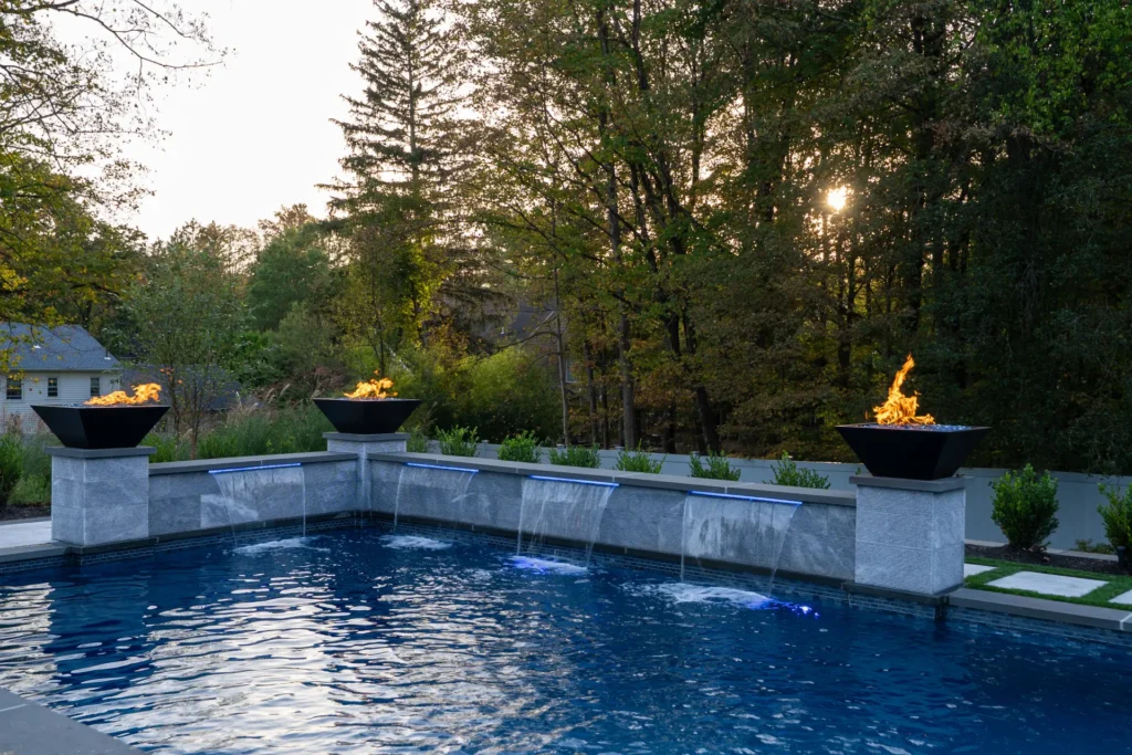 Pool wall with four waterfalls and three fire bowls against a wooded backdrop