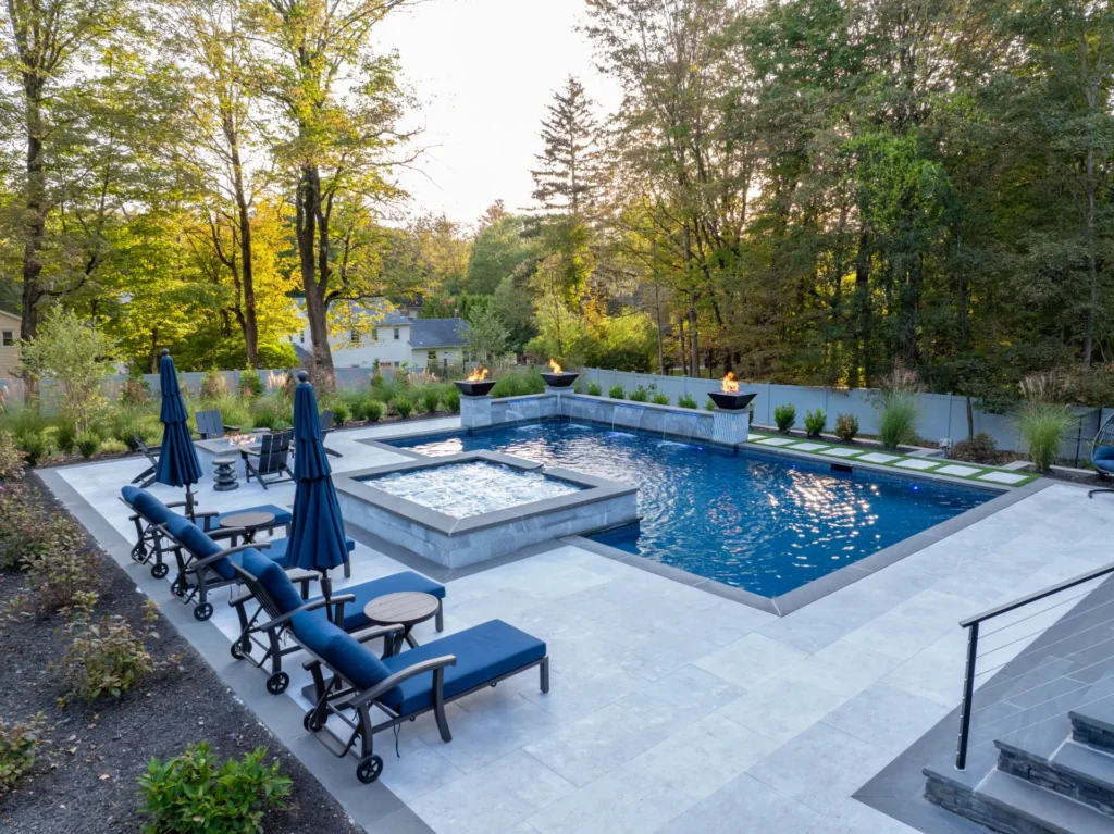 Pool and raised spa surrounded by blue loungers, fire features, and trees
